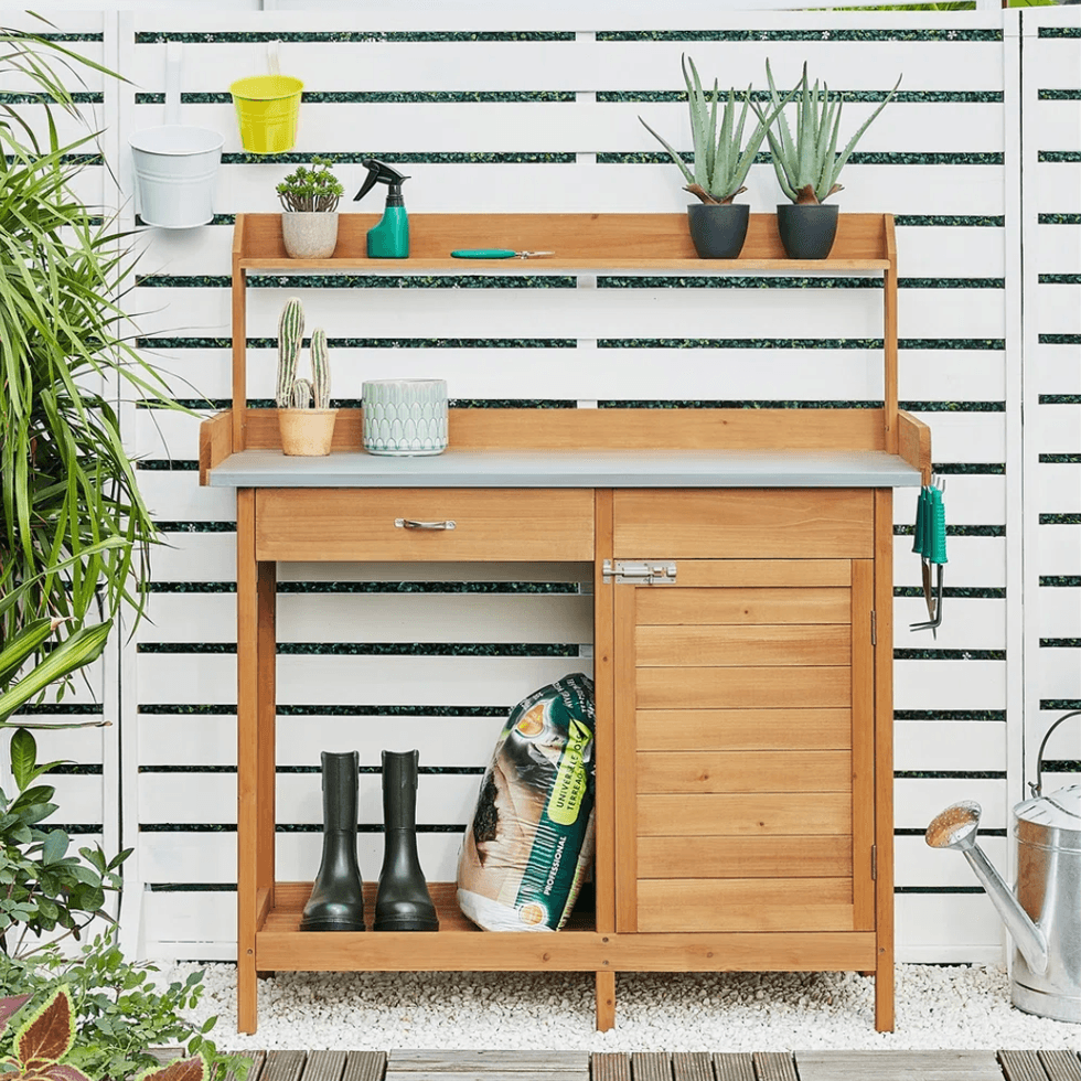 Wooden potting bench with tools, potted plants, and garden boots against a white slatted fence.