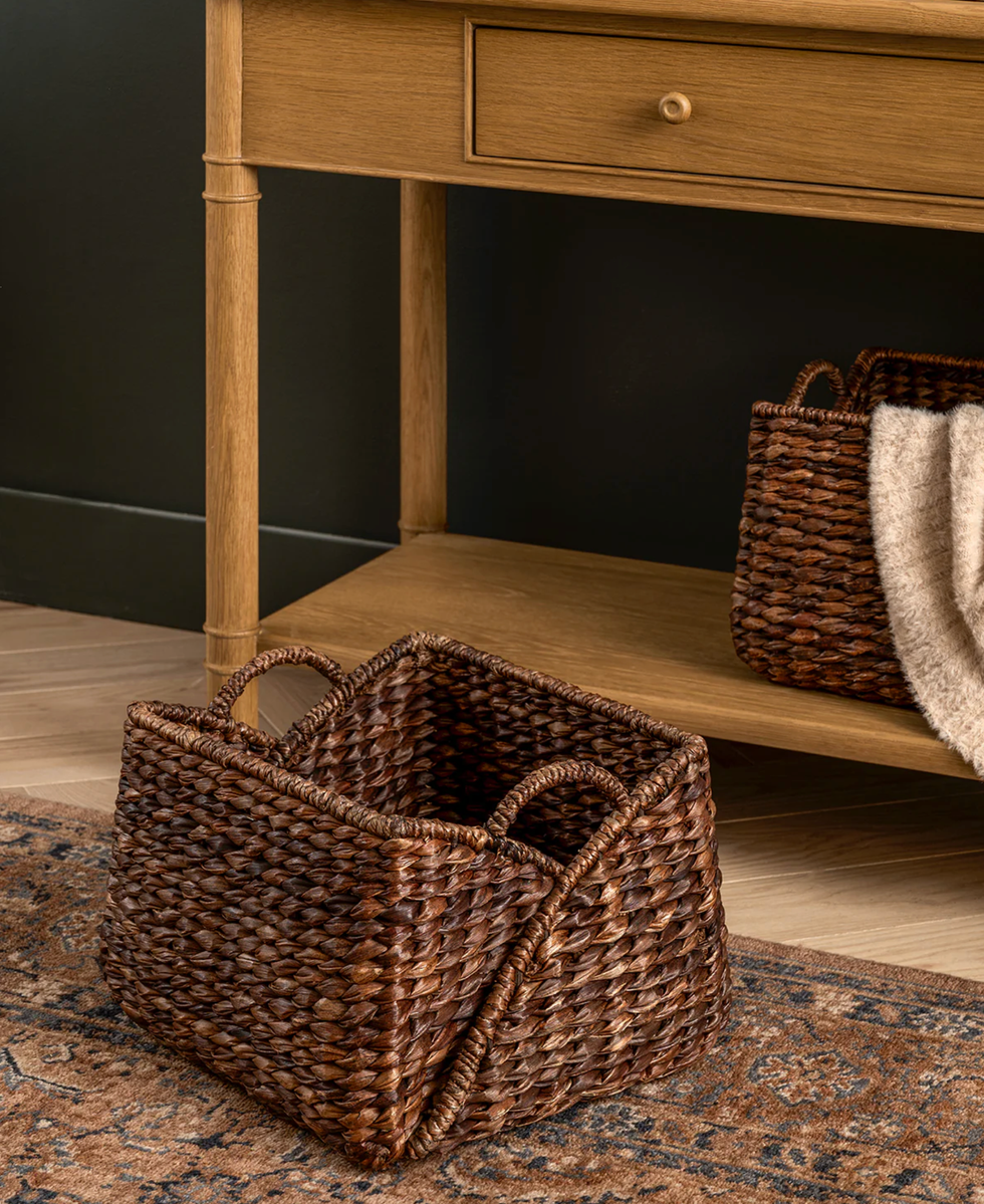 Woven basket on a patterned rug beside wooden table with drawer and basket with blanket.