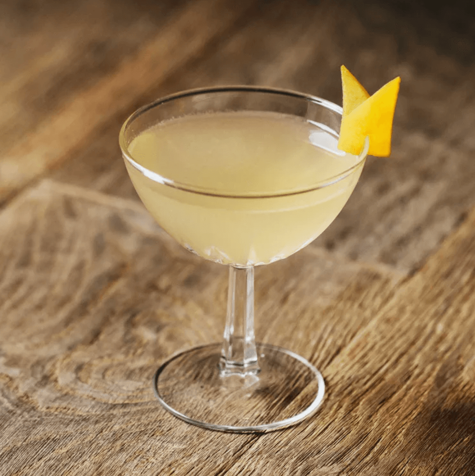 Yellow cocktail in a glass with citrus peel garnish on a wooden table.