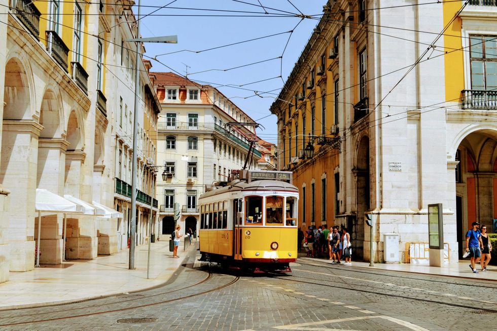Yellow tram passes through a historic European street with archways and pedestrians.