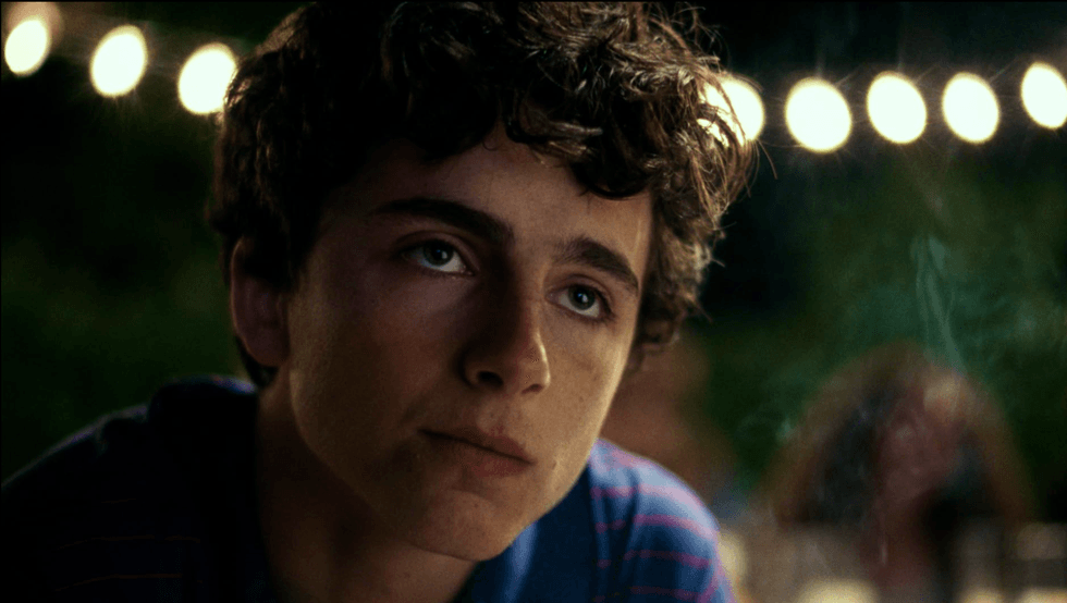 Young man with curly hair, wearing a striped shirt, gazes thoughtfully with lights behind.