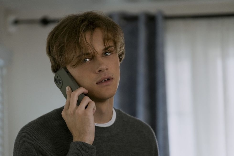 Young man with tousled hair talking on a smartphone, wearing a gray sweater indoors. chris briney as conrad
