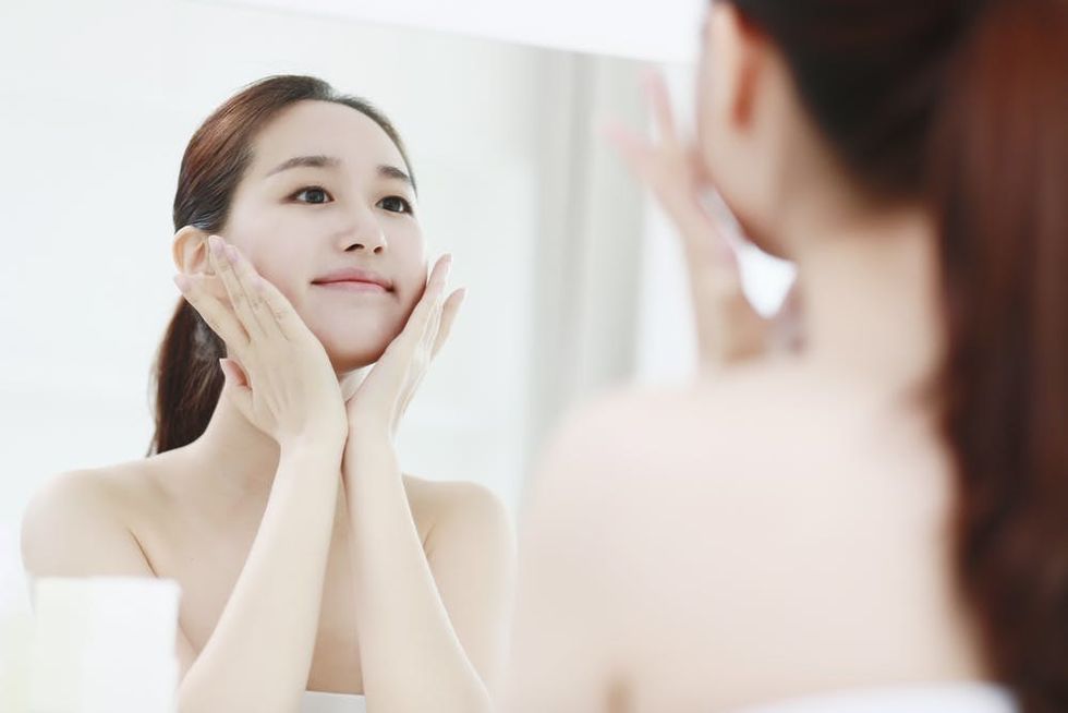 Young woman looking in the mirror while smiling at herself and holding her face in her hands.